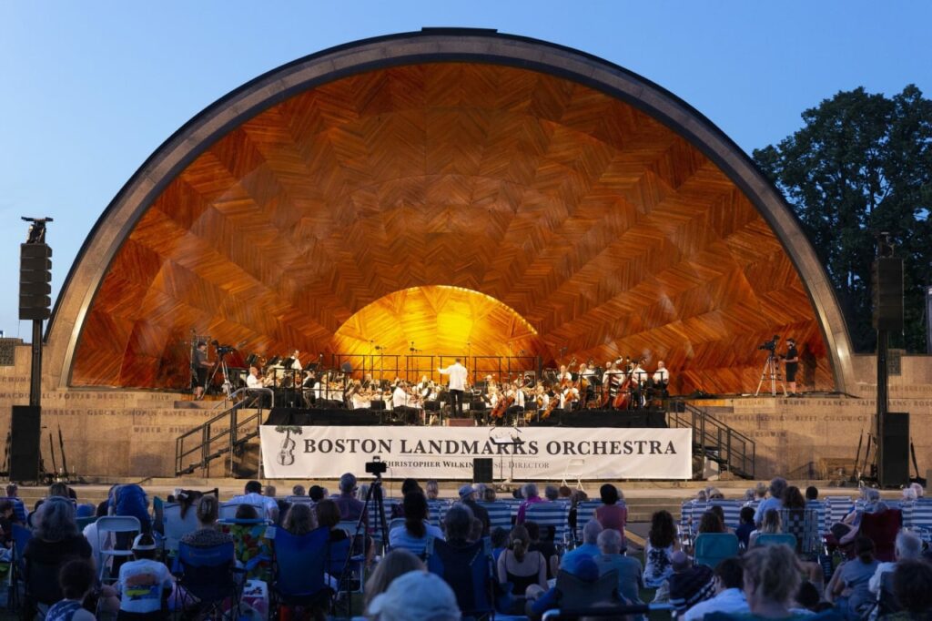 boston-landmarks-orchestra-concerts-at-the-hatch-shell The band shell with the Boston Landmarks Orchestra playing in it and an audience sitting in the grass in front of it at twilight.