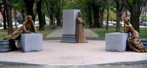 A photo of the Boston Women's Memorial - three grey columns spread out across the entrance to a walkway at the Boston Common, each with a bronze statue of a notable women from Boston history leaning or sitting on it.