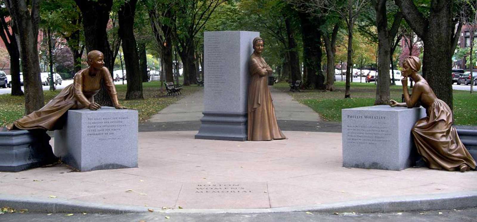 A photo of the Boston Women's Memorial - three grey columns spread out across the entrance to a walkway at the Boston Common, each with a bronze statue of a notable women from Boston history leaning or sitting on it.