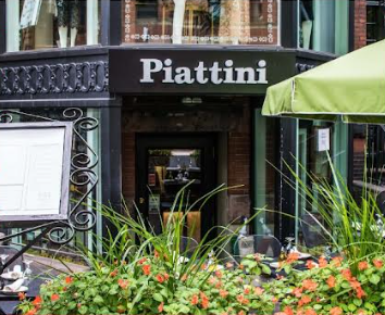 The exterior of Piattini - a restaurant in Boston. A doorway set on a corner with a black awning over it extending at an angle to the left and right of the doorway with the word "Piattini" in white letters over the doorway. In the foreground is a row of flowers with part of the menu on a stand visible on the left and part of a green outdoor table umbrella visible on the right
