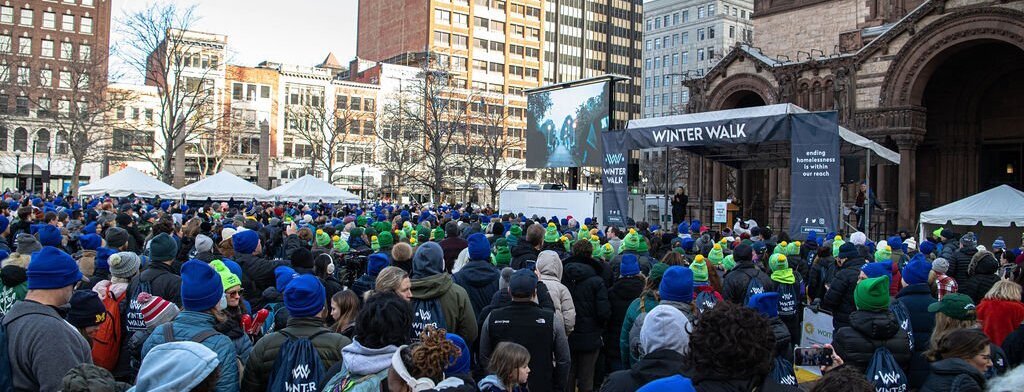Winter Walk a crowd of people gathered facing a stage with a canopy with the words "Winter Walk" on it in a square in Boston