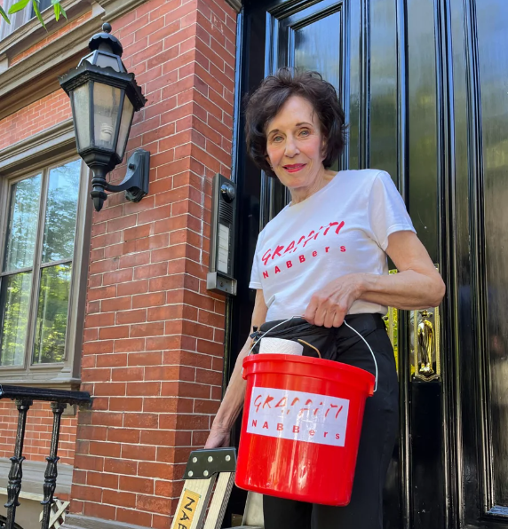 AnneSwanson-BostonComFeature Anne Swanson (a white woman with short dark brown hair) stands outside a door of a brick building in the Back Bay with a white shirt on that says "Graffiti NABBers" on it in red, holding a step stool in her right hand and a red bucket with a Graffiti NABBers sticker on it